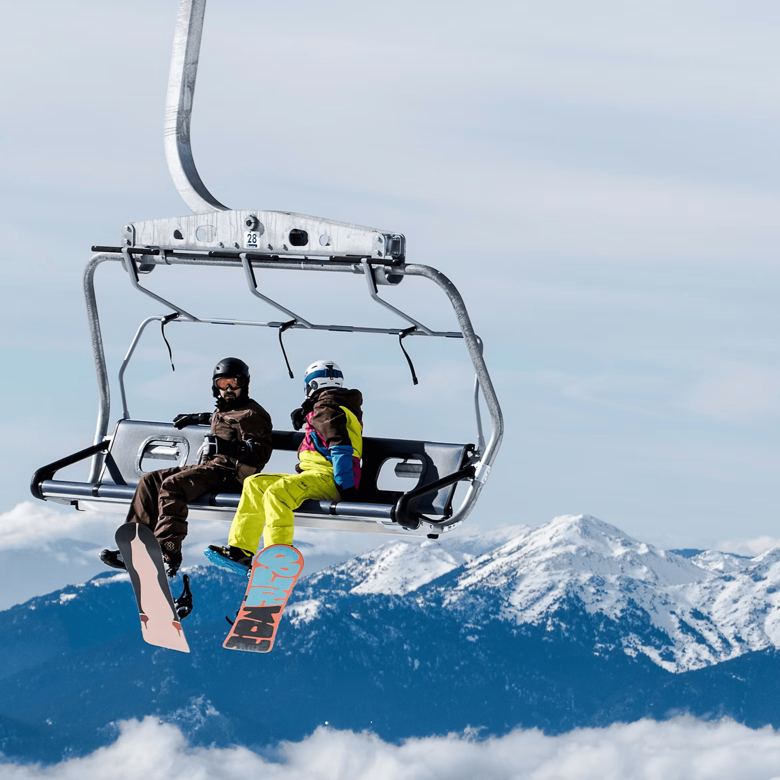 Two snowboarders on a chairlift with snow-covered mountains in the background
